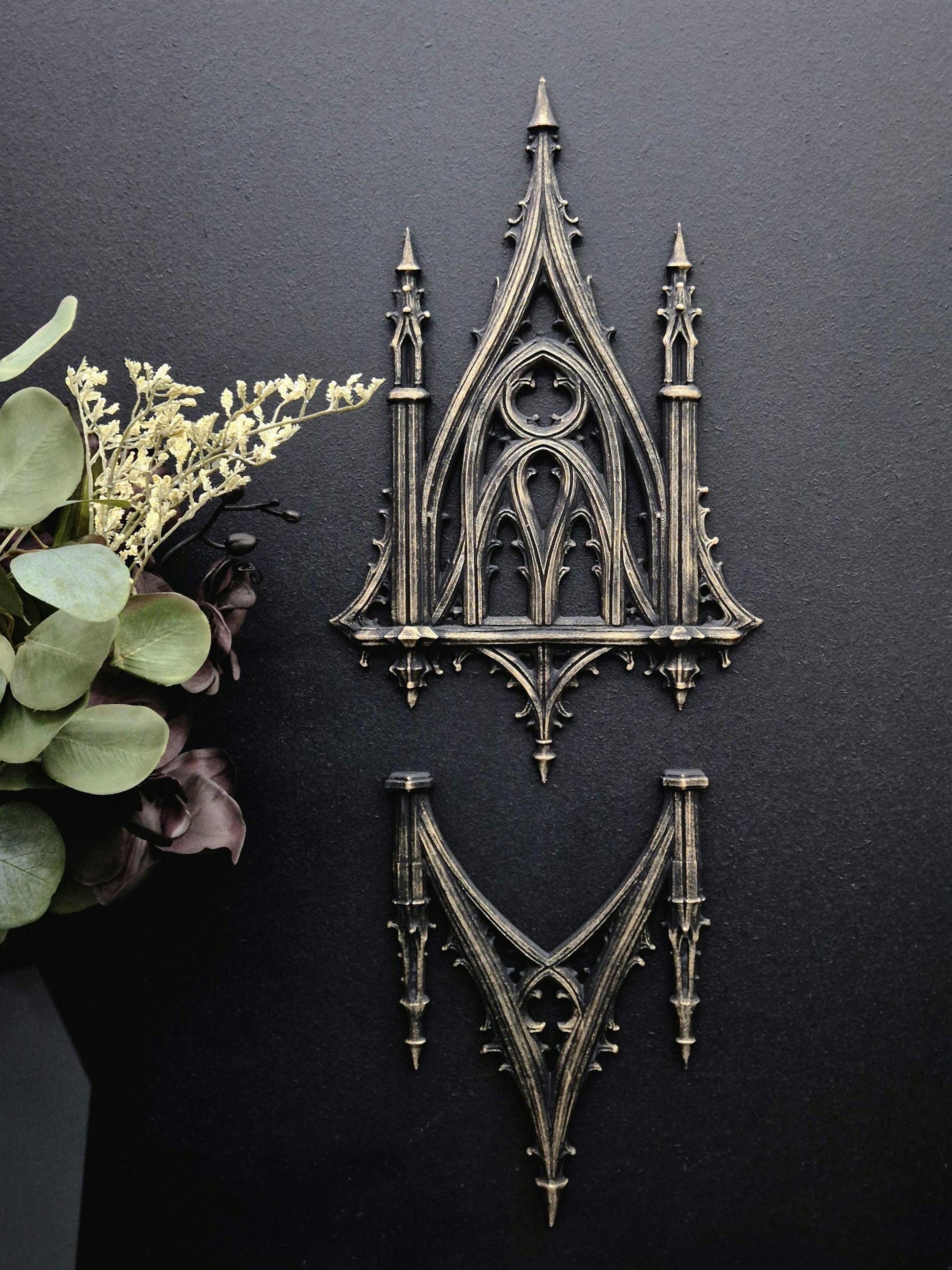 May include: Two gothic-style wall decor pieces in a dark bronze finish. The upper piece is a detailed arched design with spires, while the lower piece is a stylized V-shape. The background is a dark, textured surface.