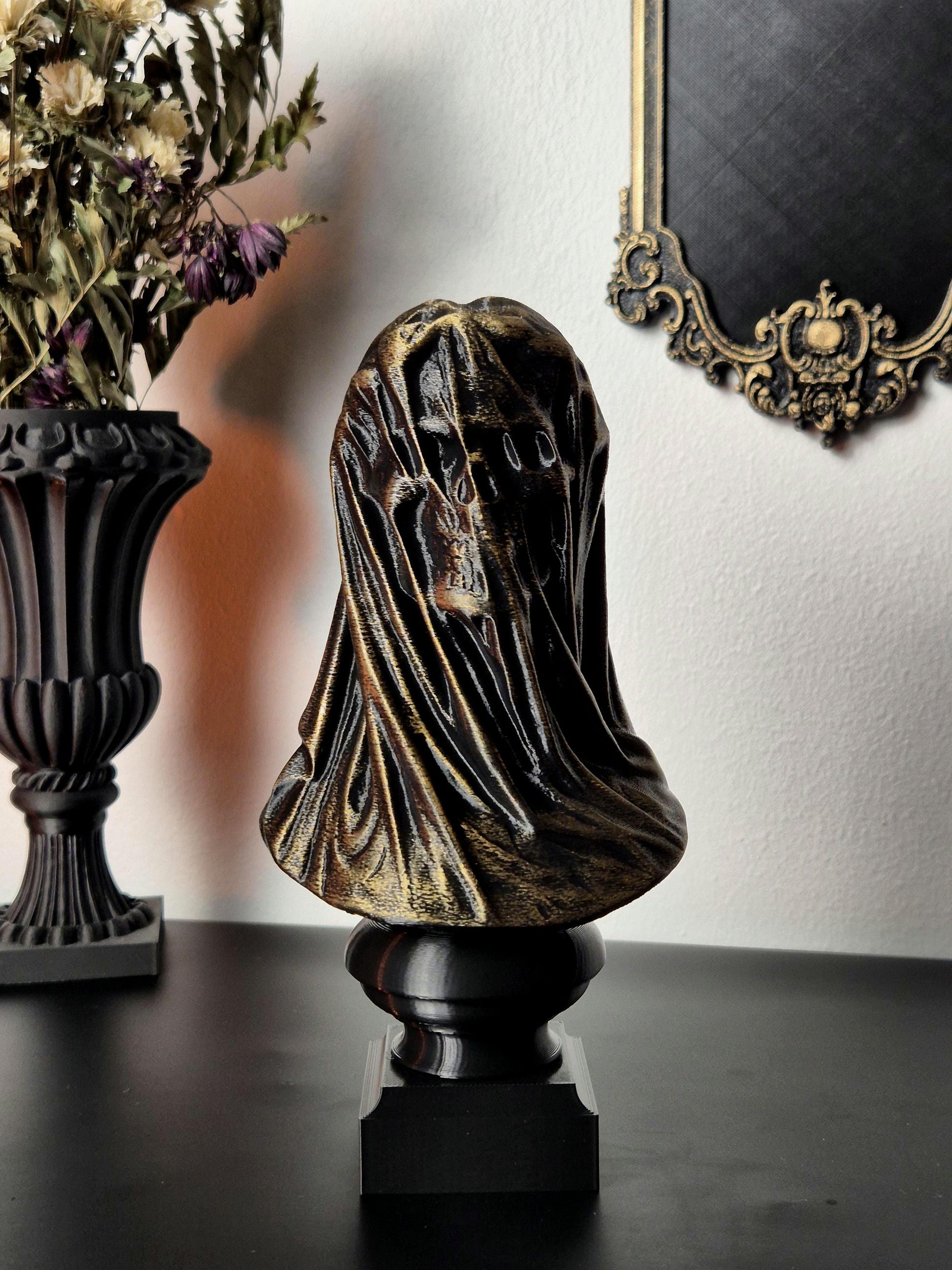 May include: A gold-painted skull bust sculpture with a draped cloth covering the head and face. The bust is on a black square base.