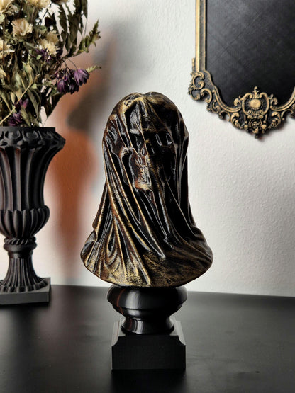 May include: A gold-painted skull bust sculpture with a draped cloth covering the head and face. The bust is on a black square base.