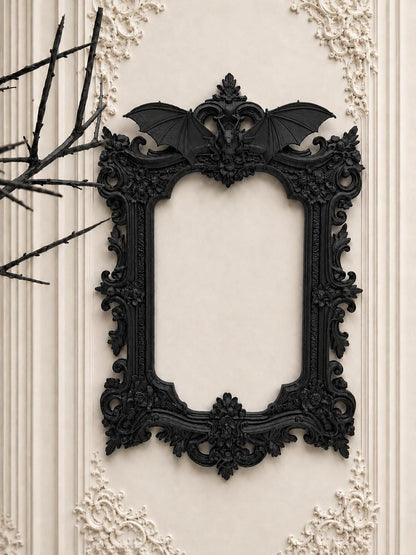 May include: Ornate black picture frame with a bat motif at the top. The frame has a detailed, gothic design with floral and scrollwork accents. The frame is mounted on a cream-colored wall with decorative molding.