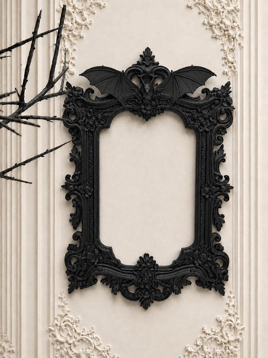 May include: Ornate black picture frame with a bat motif at the top. The frame has a detailed, gothic design with floral and scrollwork accents. The frame is mounted on a cream-colored wall with decorative molding.
