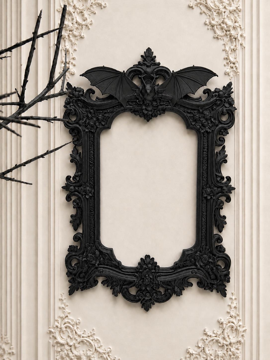 May include: Ornate black picture frame with a bat motif at the top. The frame has a detailed, gothic design with floral and scrollwork accents. The frame is mounted on a cream-colored wall with decorative molding.