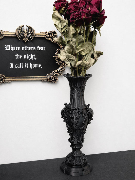 May include: A tall black vase with ornate scrollwork and a flared rim, holding a bouquet of deep red dried roses. Beside it hangs a rectangular black sign with a gold-toned frame, a bat emblem, and the text: Where others fear the night, I call it home.